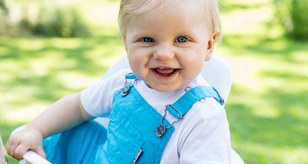 A toddler in blue overalls sits outdoors, holding a white object.