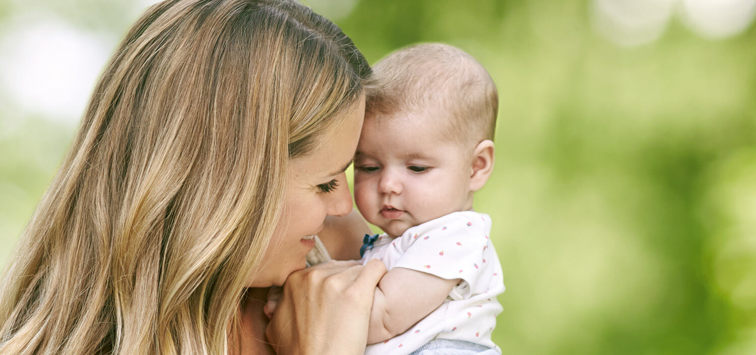 A woman with long hair holds a baby in a white outfit with red spots, both facing each other against a blurred green background.