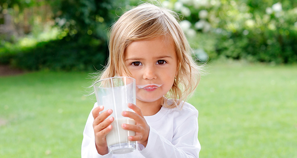 A young child with a white shirt holds a clear glass containing a white liquid, possibly milk, against a green outdoor background.