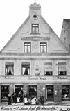 A group of people stands in front of a two-story building with a triangular roof.