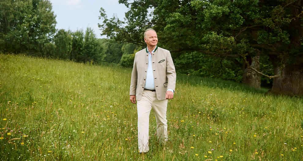 A man in a beige jacket and white pants walks through a grassy field with yellow flowers.
