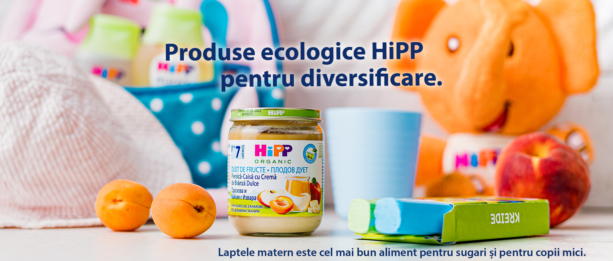 Pe o masă sunt așezate un borcan de mâncare organică pentru copii, o pălărie, un elefant de jucărie, o piersică, o cutie de creioane colorate și o ceașcă albastră.