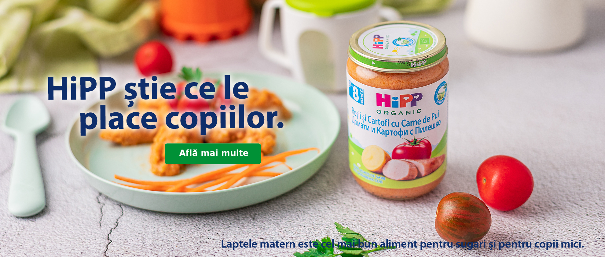 Pe o masă sunt așezate un borcan de mâncare pentru copii cu o lingură, o farfurie cu mâncare și o roșie.