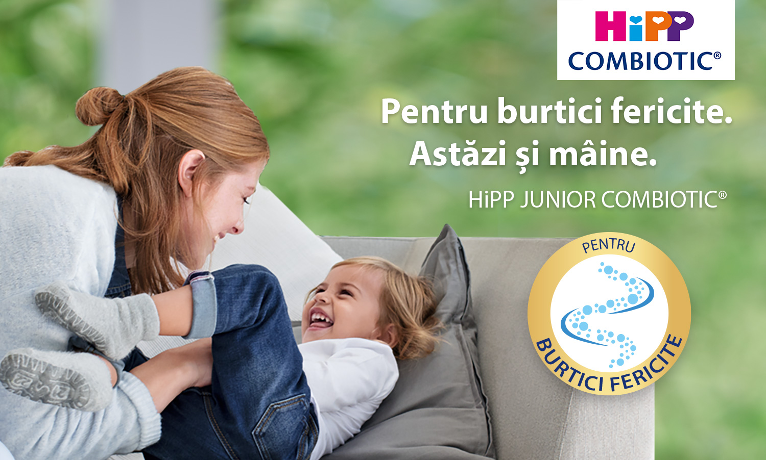 O femeie și un copil care râde sunt prezentați pe o canapea, femeia ținând picioarele copilului, iar pe fundal se află un logo și un text care promovează un produs numit "HiPP JUNIOR COMBIOTIC®".
