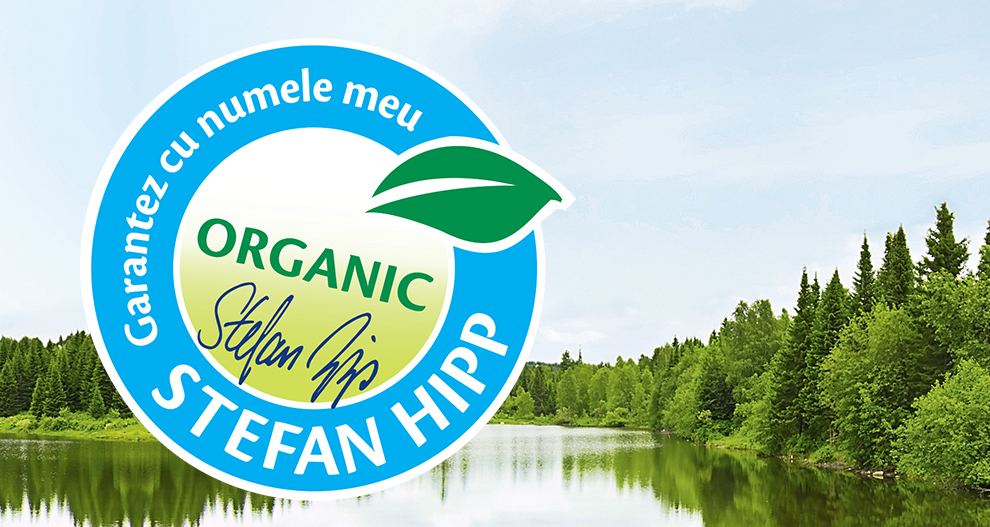 Un peisaj senin al unui lac înconjurat de copaci verzi și dese este suprapus cu un logo circular albastru care prezintă cuvântul "ORGANIC" și numele "Stefan Hipp".