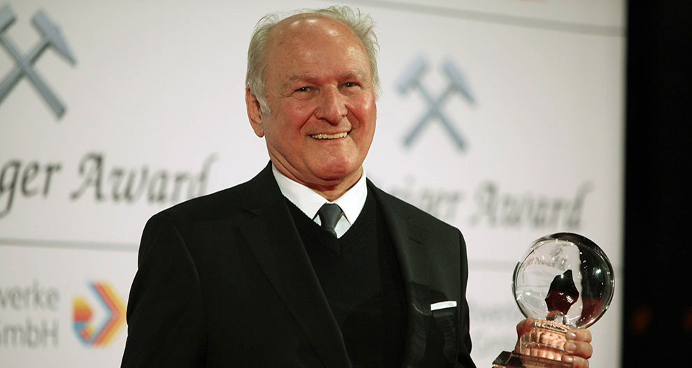 Un bărbat în vârstă în costum negru stă pe o scenă, ținând în mână un premiu sferic transparent, cu cuvintele "Schweizer Award" și logo-uri în fundal.