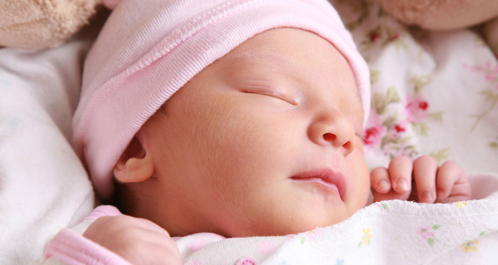 Un prim plan al feței unui bebeluș care doarme, purtând o căciuliță roz și înfășurat într-o pătură albă cu motive florale.