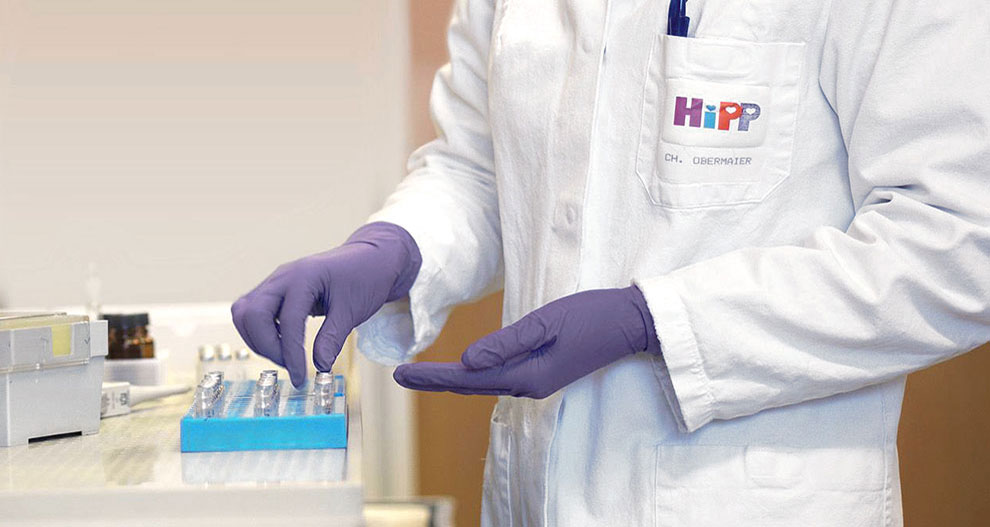 O persoană în halat de laborator alb cu sigla "HiPP" și numele "CH. OBERMAIER" poartă mănuși mov și manipulează o tavă albastră cu mai multe recipiente mici.