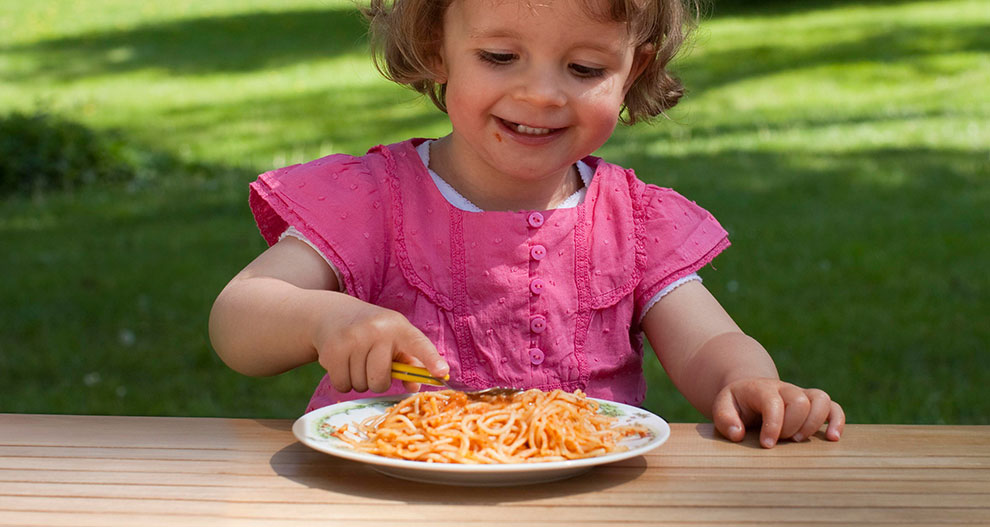 Un copil în tricou roz mănâncă spaghete dintr-o farfurie albă.