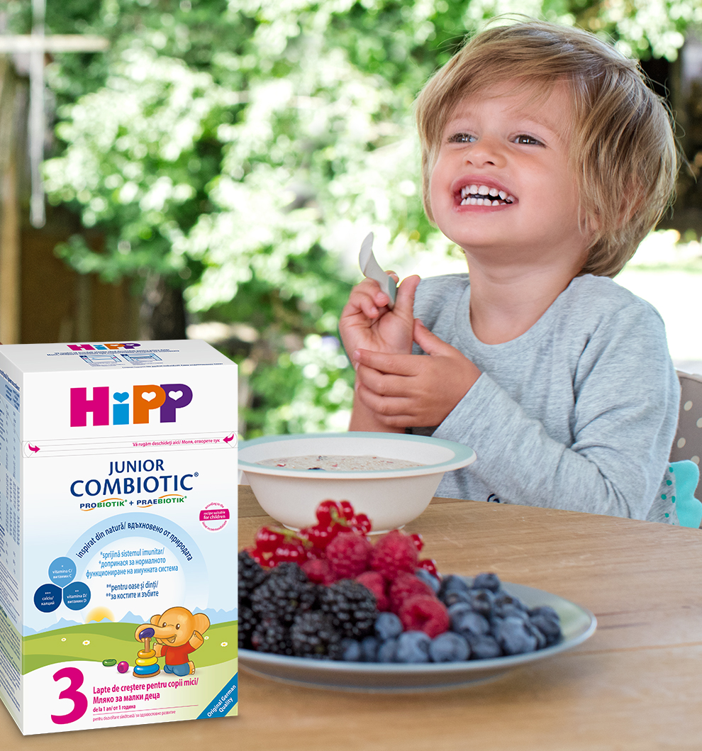 Un copil mic este așezat la o masă cu un bol de mâncare și o farfurie cu fructe de pădure, lângă o cutie de "HiPP Junior Combiotic" cu o ilustrație de desene animate.