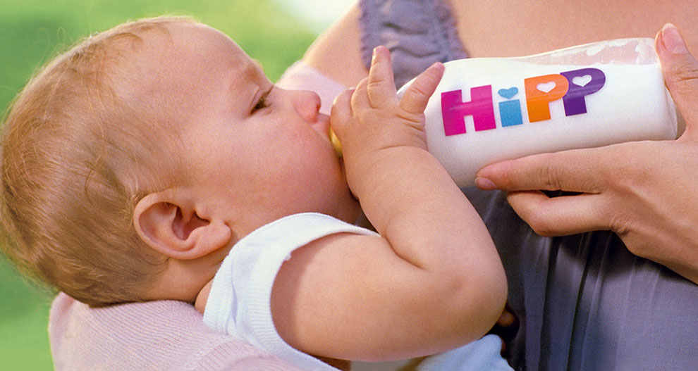 Un bebeluș bea dintr-un biberon pe care scrie "HiPP".