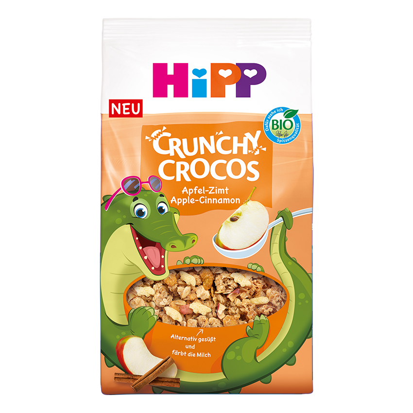 Un pachet de cereale Crunchy Crocos cu un personaj crocodil verde, o felie de măr și un baton de scorțișoară, însoțit de un text în limba germană.